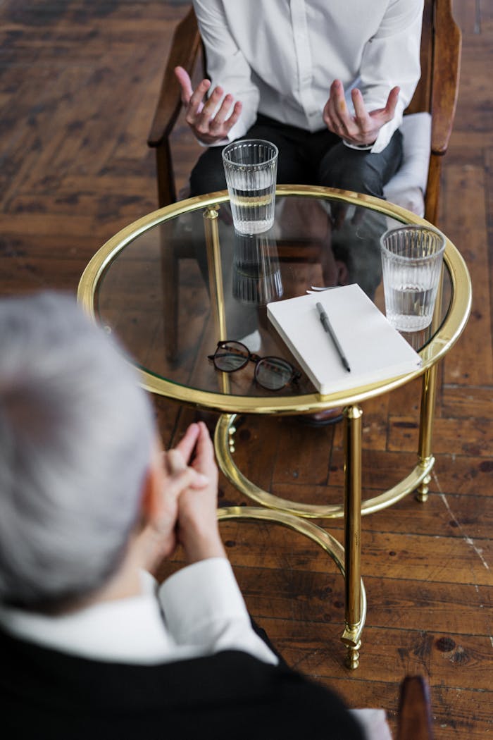 our-services-1 Two adults discussing mental health in a counseling session across a glass table indoors.